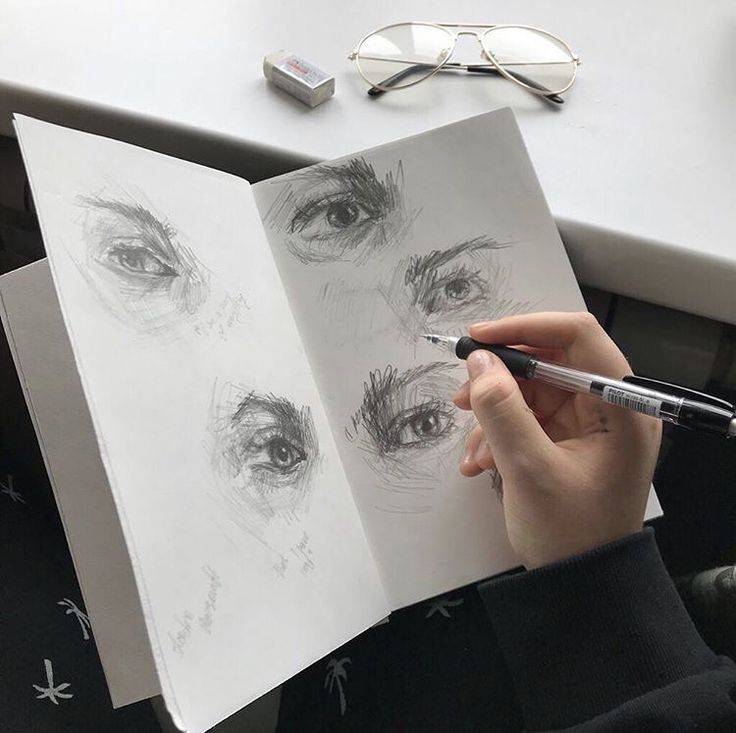 a person holding a pen and drawing on paper with eyeballs in the background next to a pair of glasses