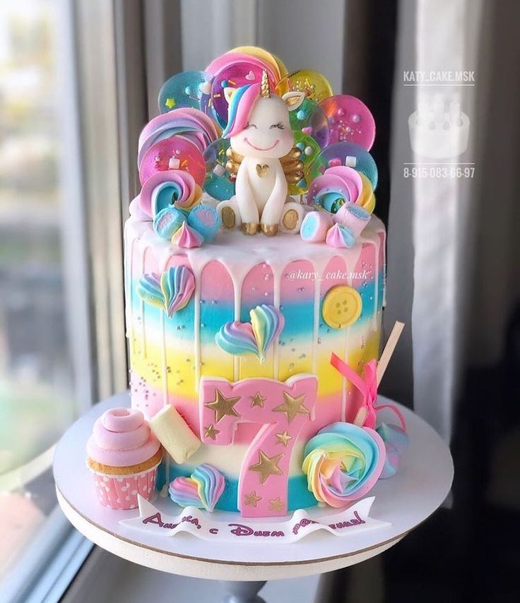 a birthday cake decorated with unicorns and rainbow candies is on a table near a window