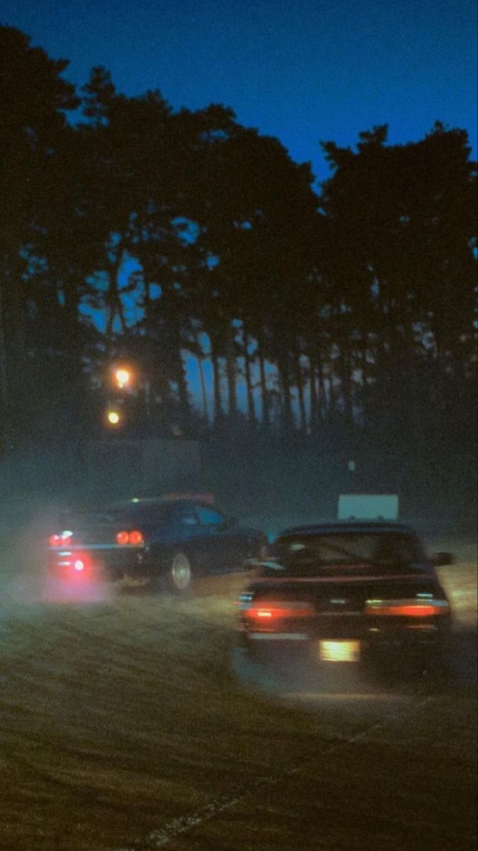 two cars driving down a road at night with the headlights on and trees in the background