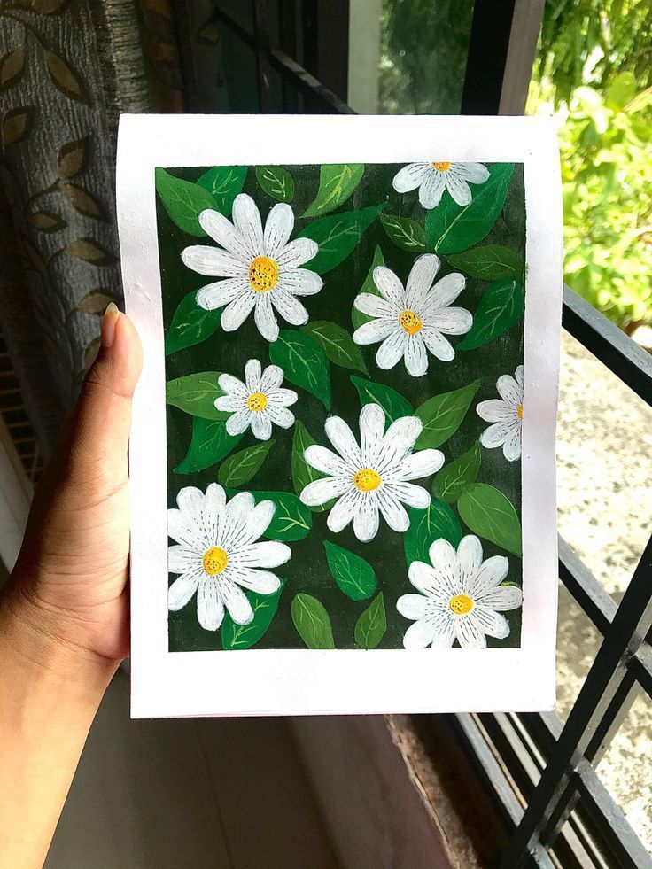 a hand holding up a card with white daisies on green leaves in front of a window