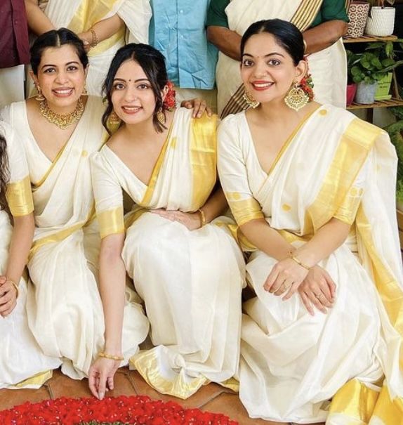 four women in white and yellow saris posing for a photo with flowers on the floor