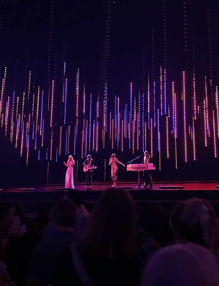 three people standing on stage in front of an audience at a musical performance with colorful lights behind them