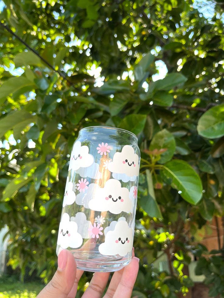 someone holding up a glass with clouds and flowers painted on the side, in front of a tree