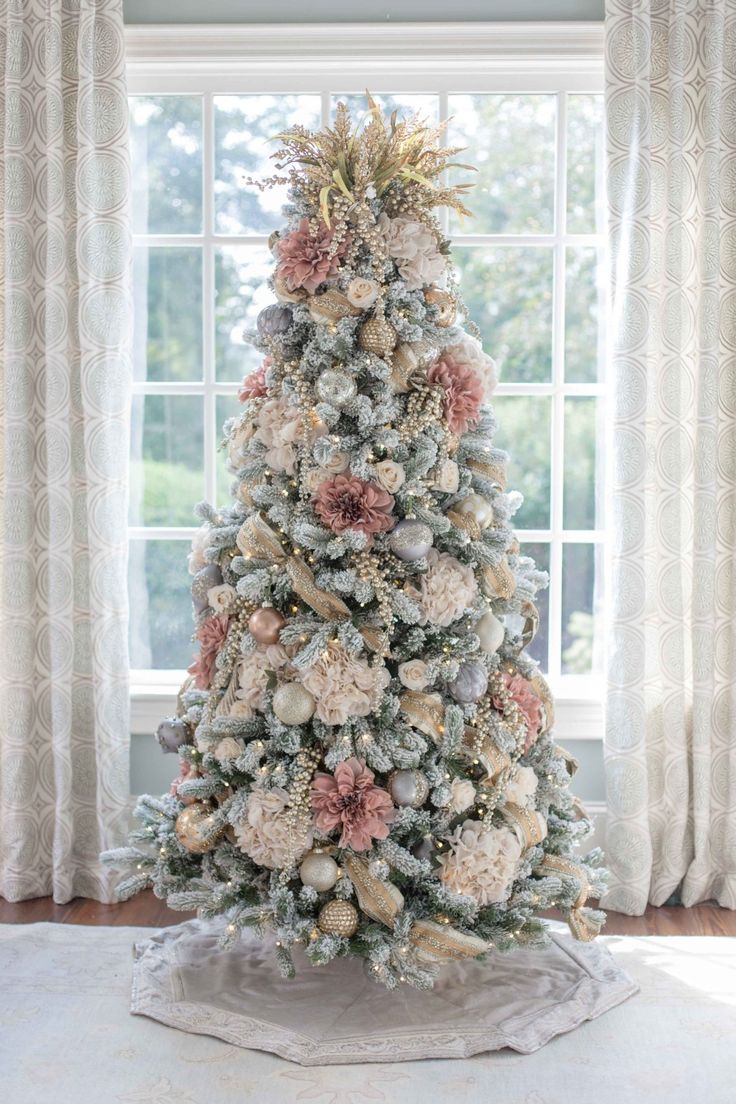 a decorated christmas tree in front of a window with white drapes and pink flowers