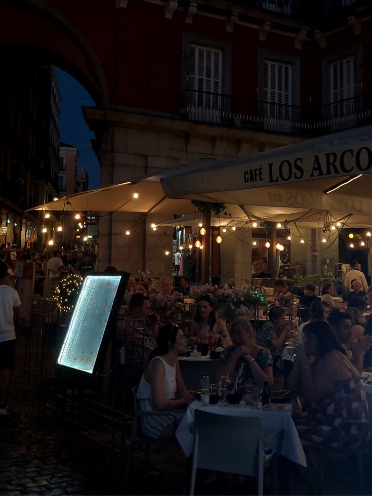 people are sitting at tables in front of an outdoor restaurant with lights on the roof