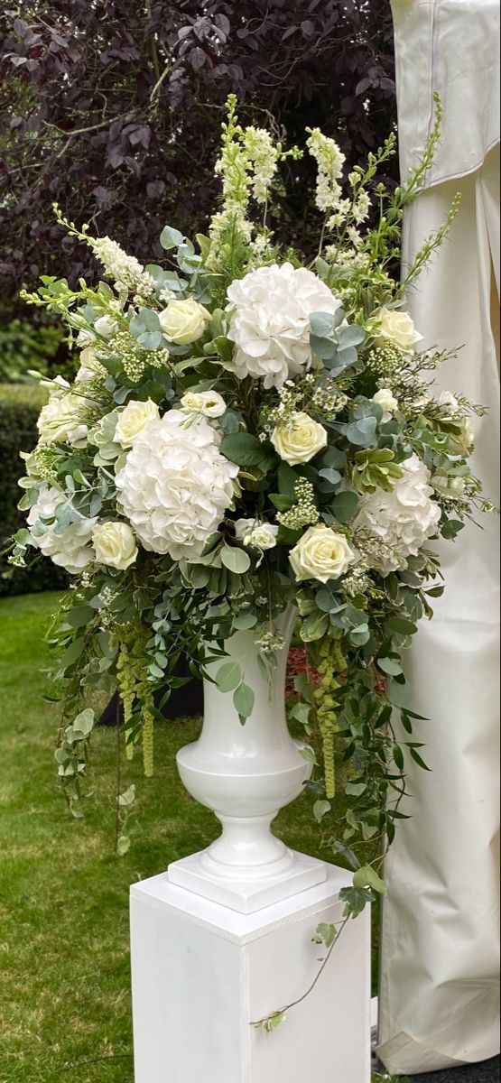 a white vase filled with flowers sitting on top of a grass covered field next to trees