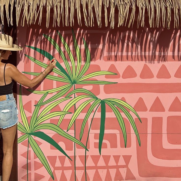 a woman standing next to a pink wall with a palm tree painted on it