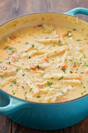 a blue pot filled with chicken and pasta soup on top of a wooden table next to a spoon