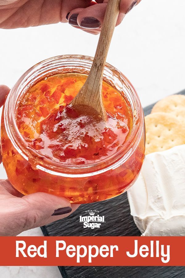 a hand holding a wooden spoon over a jar of red pepper jelly with crackers in the background