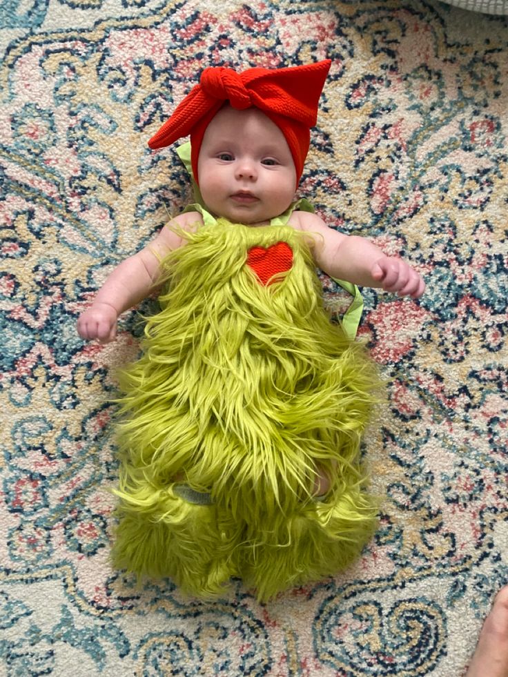 a baby is dressed up in a green outfit and red hat while laying on a rug