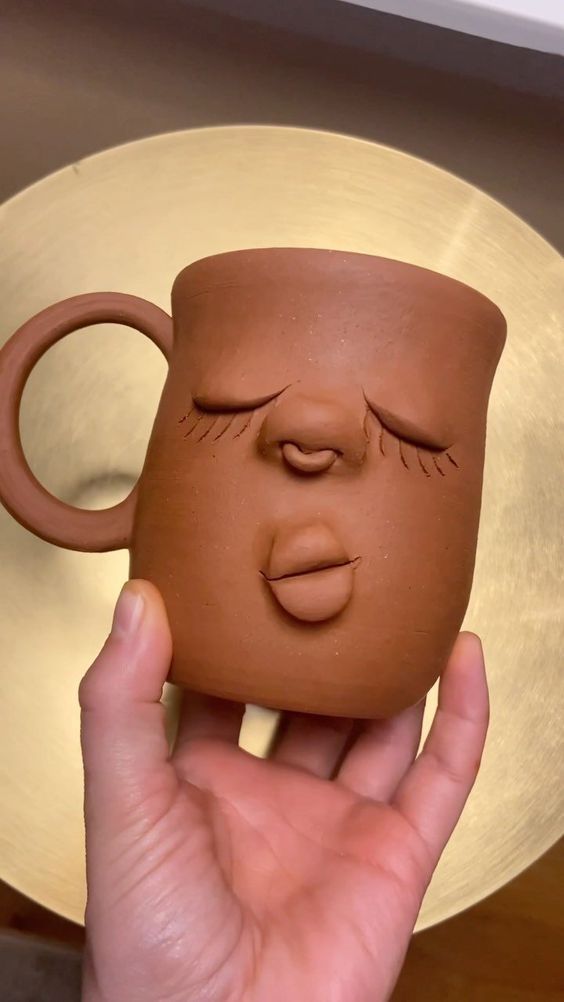 a person holding up a clay mug with their face drawn on it's side