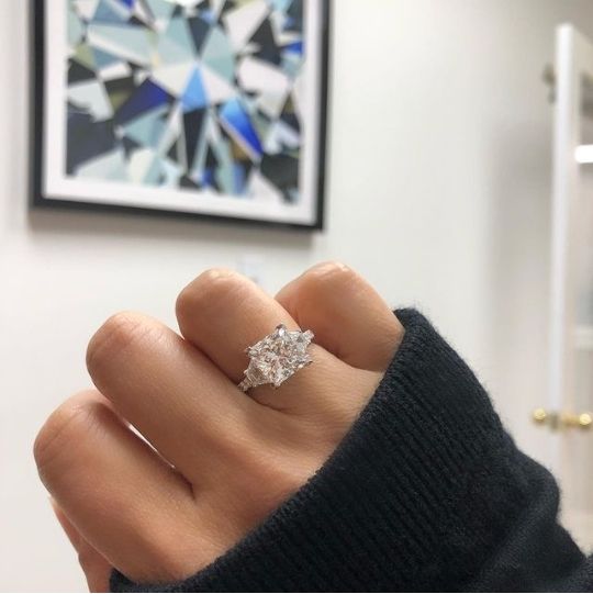 a woman's hand with a diamond ring on top of her finger in front of a painting