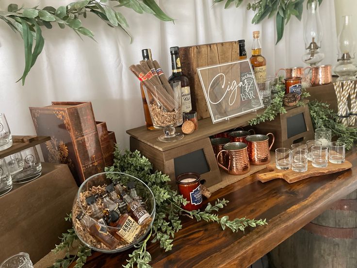 an assortment of liquor bottles and glasses on a wooden table with greenery around it