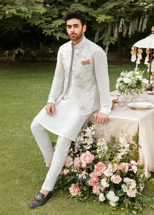 a man sitting on top of a table with flowers in front of him wearing a white suit