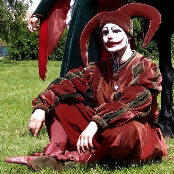 a man dressed as a jester sitting on the ground next to a black horse