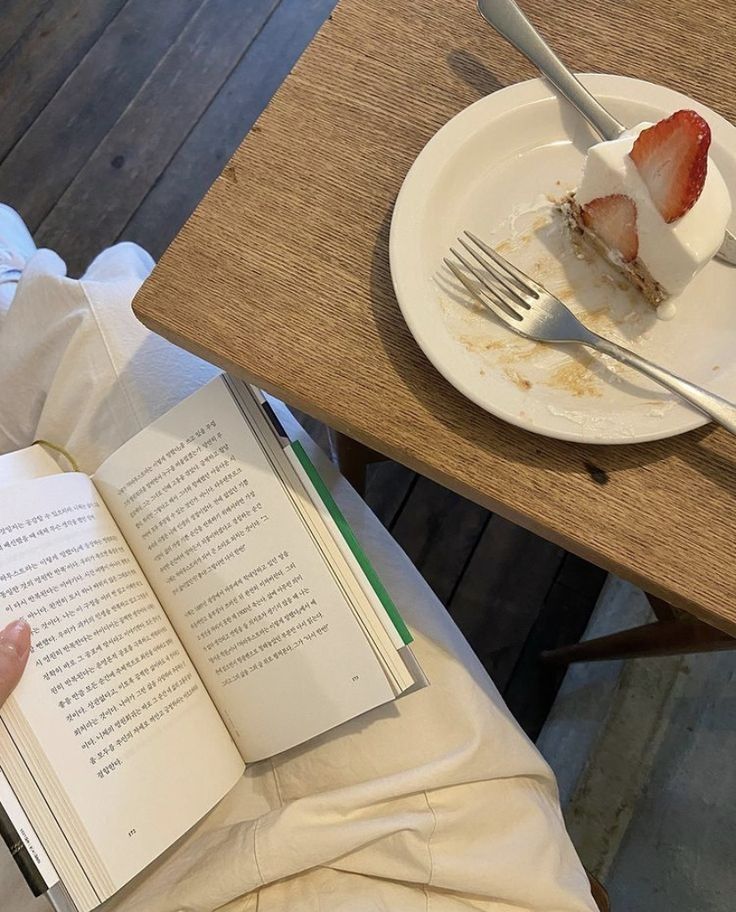 a person sitting at a table reading a book with a slice of cake on it