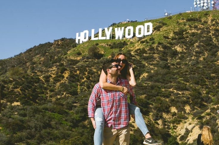 a man and woman standing in front of the hollywood sign on top of a hill