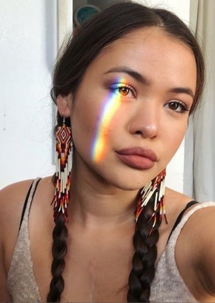 a woman with long braids and rainbow makeup looks at the camera while wearing earrings