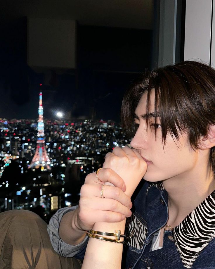 a young man sitting in front of a window with the city lights behind him at night