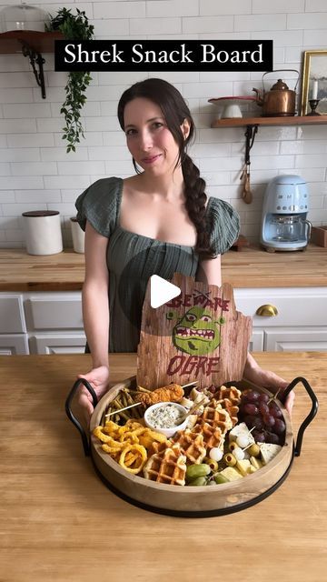 a woman sitting at a table with a plate of food in front of her and the words shrek snack board on it