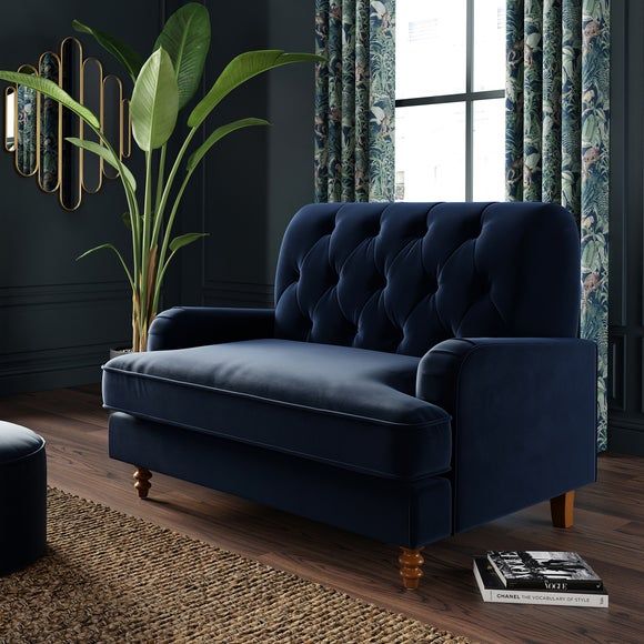 a living room with a blue velvet couch and large potted plant in the corner