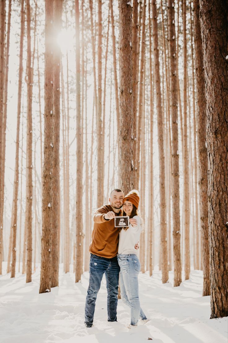 a man and woman are standing in the snow by some trees with their arms around each other