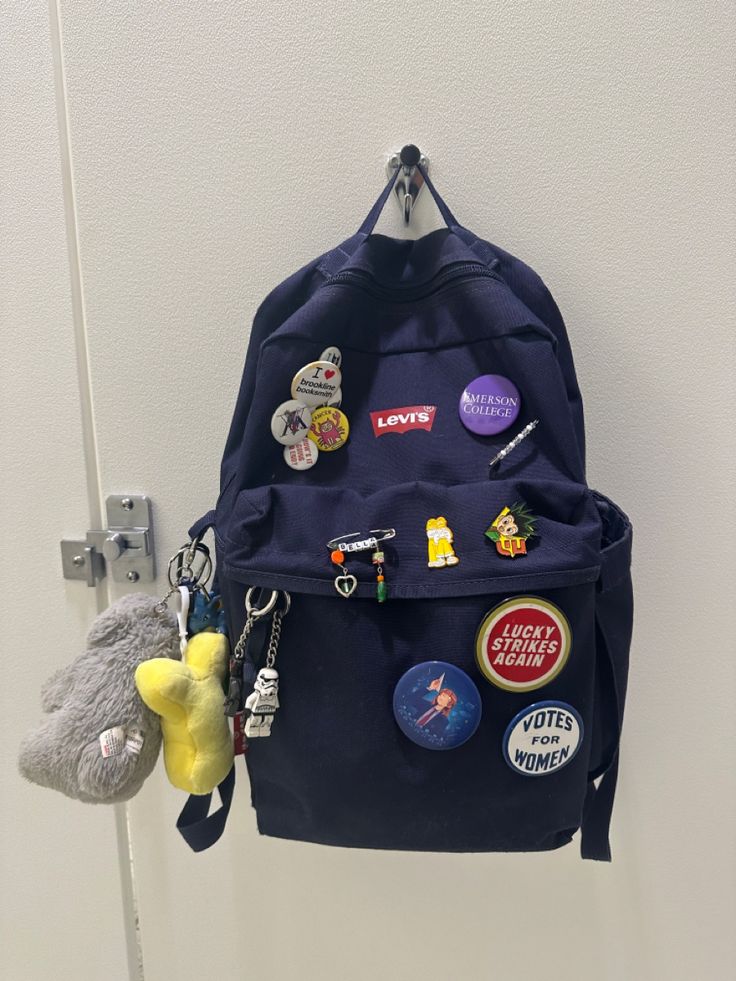 Navy blue backpack with various pins and keychains Keychains On Backpack, Backpack With Pins Aesthetic, Backpack Customize Ideas, Backpack Accessories Keychain, Levis Backpack, Grey Jansport Backpack, School Bag Keychain, Decorated Backpack, Customized Backpack