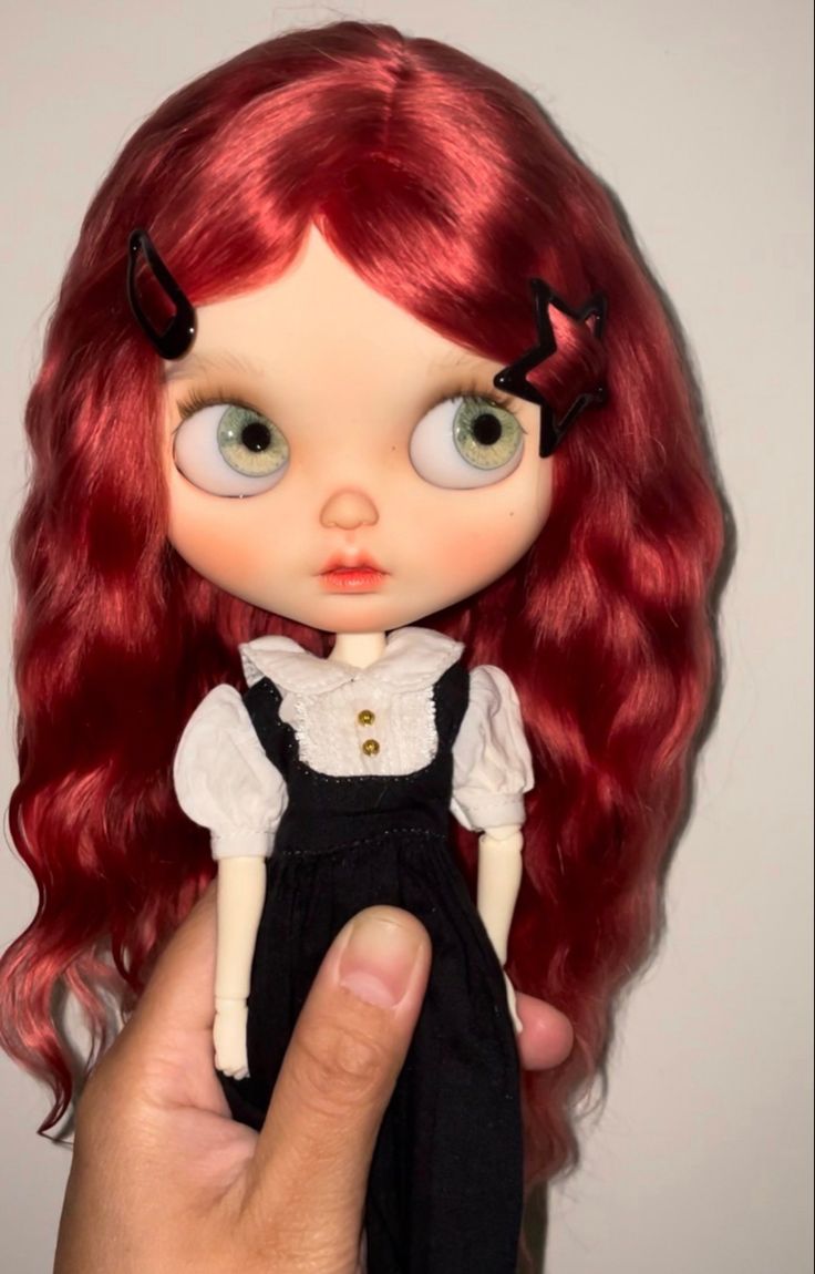 a hand holding a doll with long red hair and big eyes, wearing a black dress