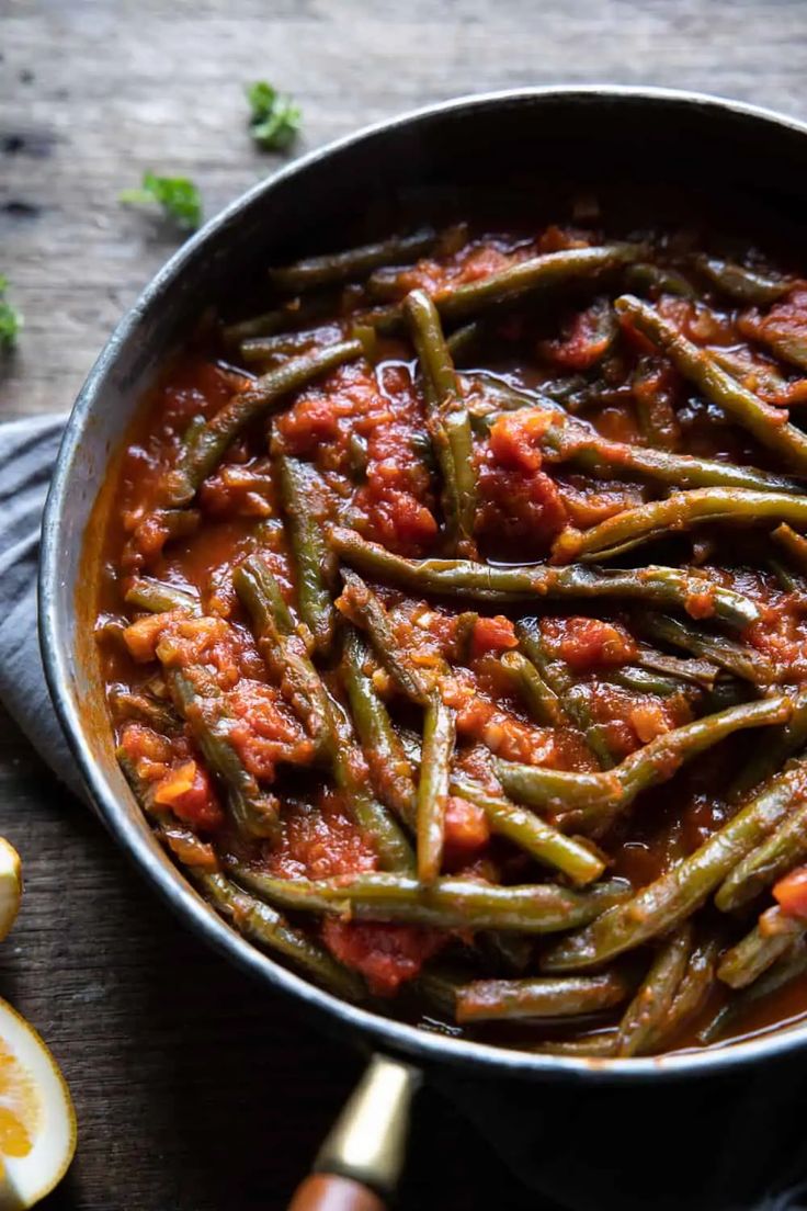 green beans with tomato sauce in a pan next to sliced lemons and parsley