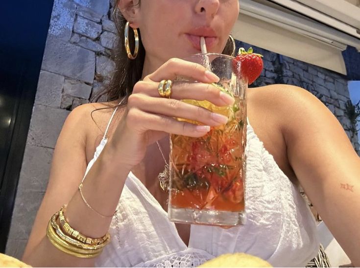 a woman drinking from a tall glass with strawberries in it and holding a straw