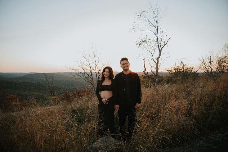 a man and woman standing on top of a hill next to each other with trees in the background