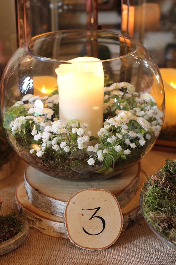 a candle is lit in a glass bowl filled with flowers and greenery on a table