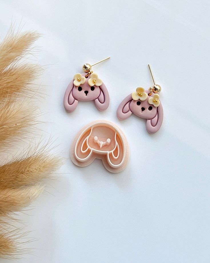 two pink earrings with ears and flowers on them sitting next to some brown feathers,