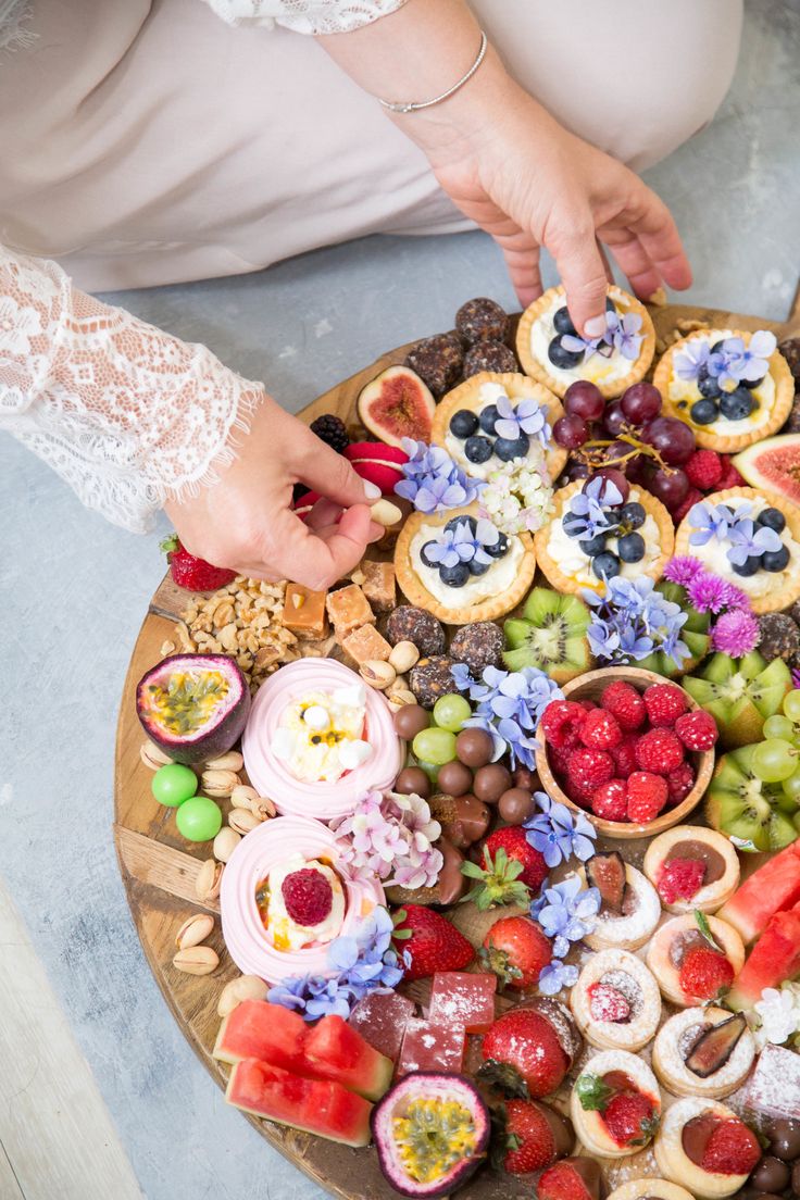 a platter filled with fruit and pastries on top of a wooden table next to a woman's hand