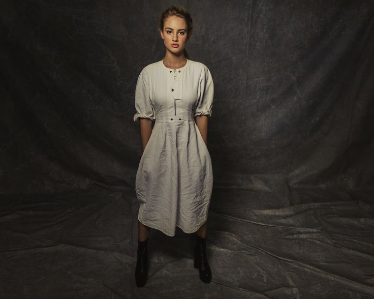 a woman standing in front of a black backdrop wearing a white dress and brown boots