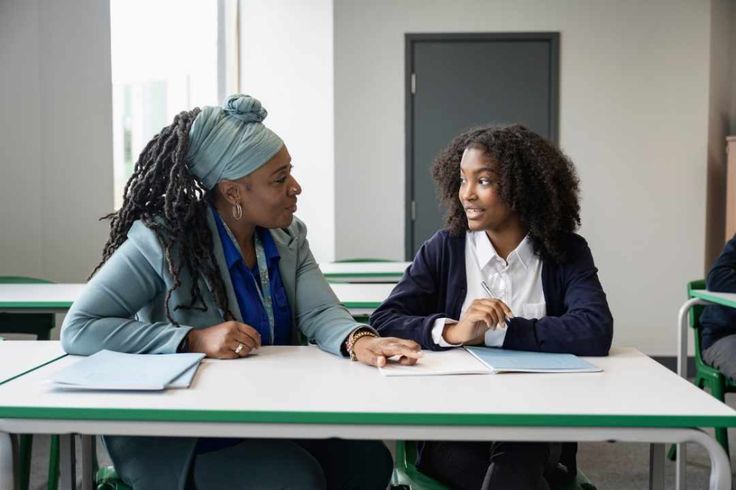 two women sitting at a table talking to each other in front of some desks