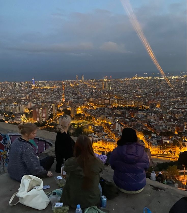 people are sitting on the edge of a hill looking at the city lights and planes in the sky