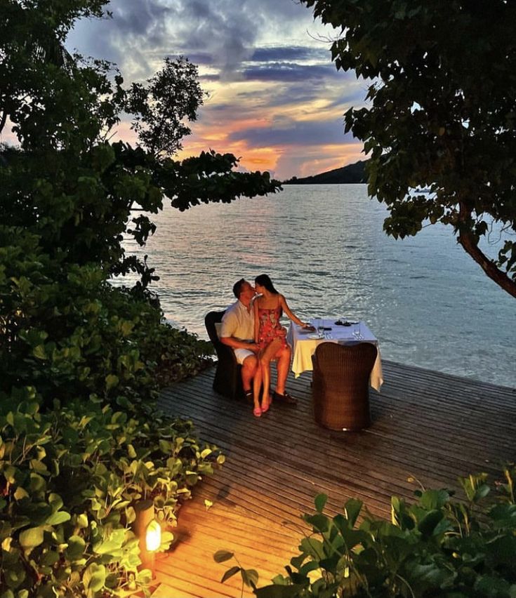 two people are sitting on a bench near the water at sunset, with candles lit in front of them