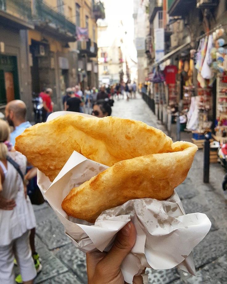 a person holding up some food in their hand on the street with people walking by