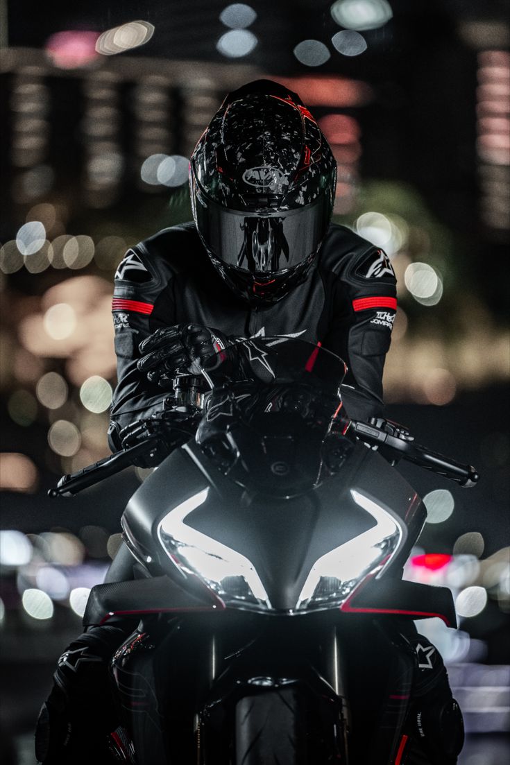 a man riding on the back of a black motorcycle at night with lights in the background