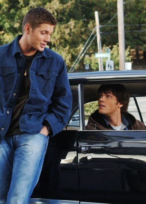 two young men standing next to each other in front of a black car and talking