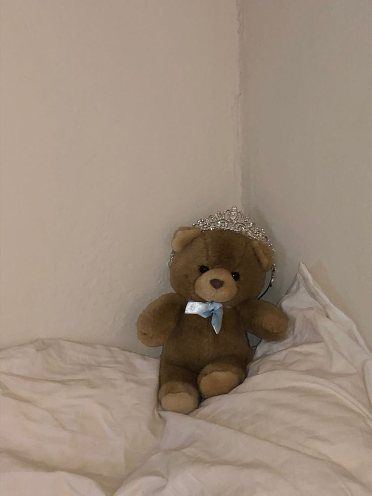 a brown teddy bear wearing a tiara sitting on a white comforter in a corner