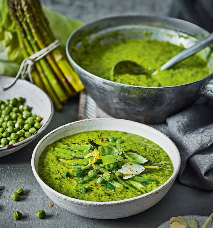 two bowls filled with green pea soup next to asparagus and other vegetables on a table