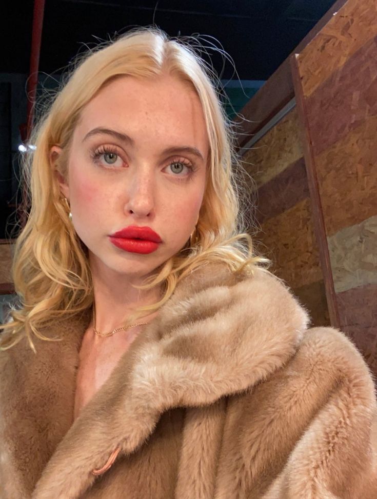 a woman with blonde hair wearing a fur coat and red lipstick in front of a wooden wall