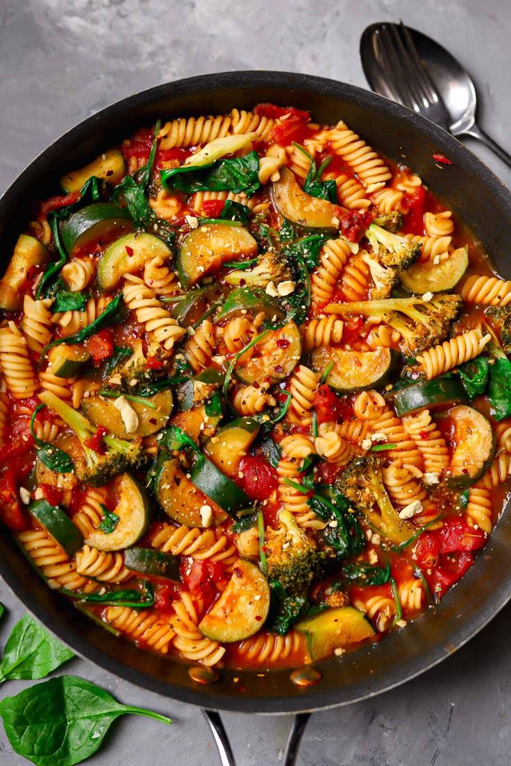 a skillet filled with pasta and vegetables on top of a gray table next to silverware