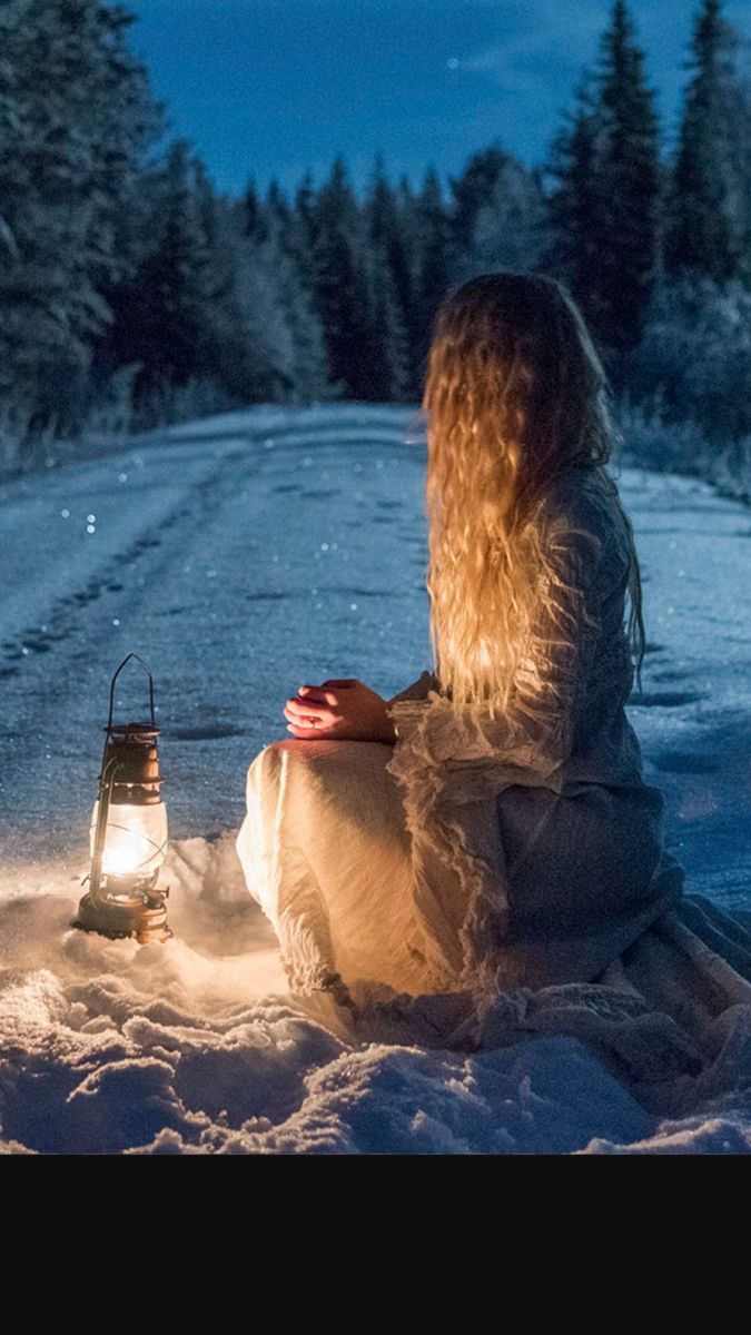 a woman sitting in the snow next to a lantern on a snowy road at night