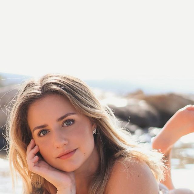 a beautiful young woman laying on top of a beach next to the ocean with her hand under her chin