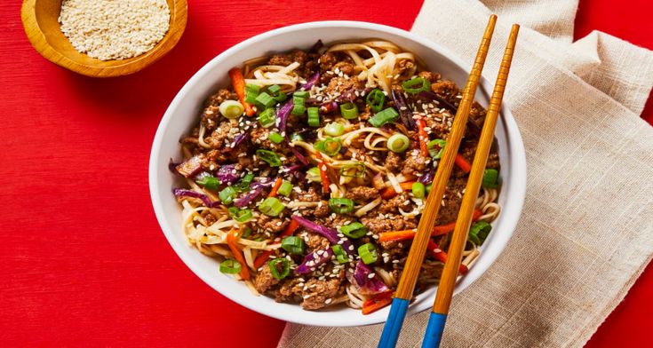 a bowl filled with noodles and vegetables next to chopsticks on a red surface