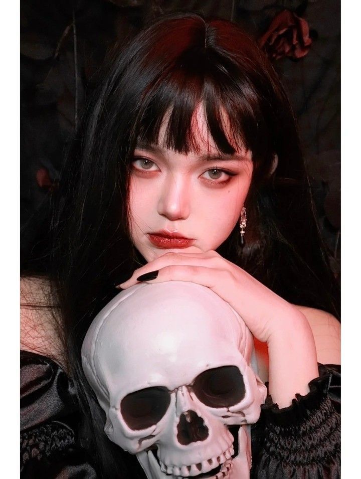 a woman with long black hair holding a skull in front of her face and looking at the camera