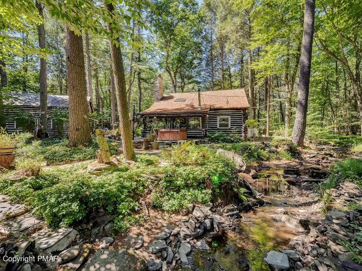 a log cabin in the woods surrounded by trees and rocks with a creek running through it